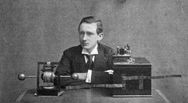 Publicity photo in a magazine of British-Italian radio entrepreneur Guglielmo Marconi with his early wireless radiotelegraphy transmitter and receiver, invented around 1895. On the left is his spark gap transmitter, consisting of a dipole antenna made of two brass rods with a spark gap between them, which transmits pulses of radio waves spelling out text messages in Morse code. On the right is his receiver, consisting of a coherer which rings a bell when it receives the dots and dashes of Morse code The bell and batteries are in the box. (Photo: Wikimedia)