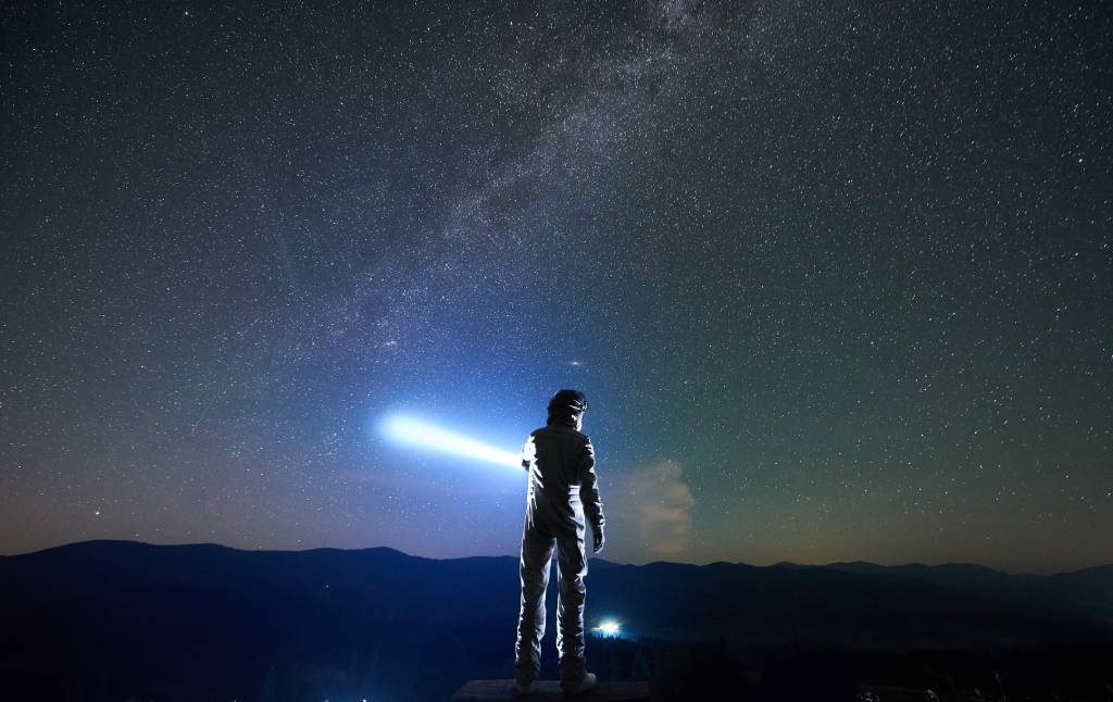 Cosmonaut wearing white space suit and helmet directs a blue ray of light into starry sky above horizon in the night, standing with his back to the camera in the mountains. (Image: Shutterstock/Anatoliy Gleb)