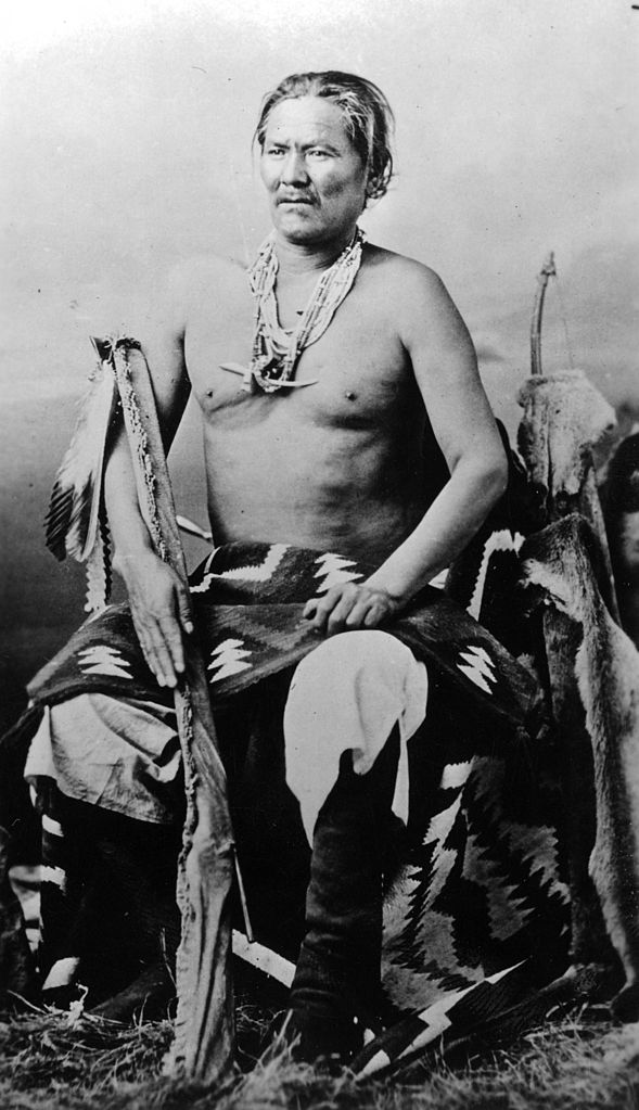 Studio portrait (sitting) of Chief Manuelito (1818–1893), a Native American (Navajo) man. He holds a rifle in a buckskin scabbard and wears moccasin boots, a blanket, and bead necklaces with a saber pendant. "Manuelito, the once fierce chief of the Navajo" and "Saved Navajo Bill's Life Once" hand-written on back of print. (Photo: Wikimedia/Denver Library)