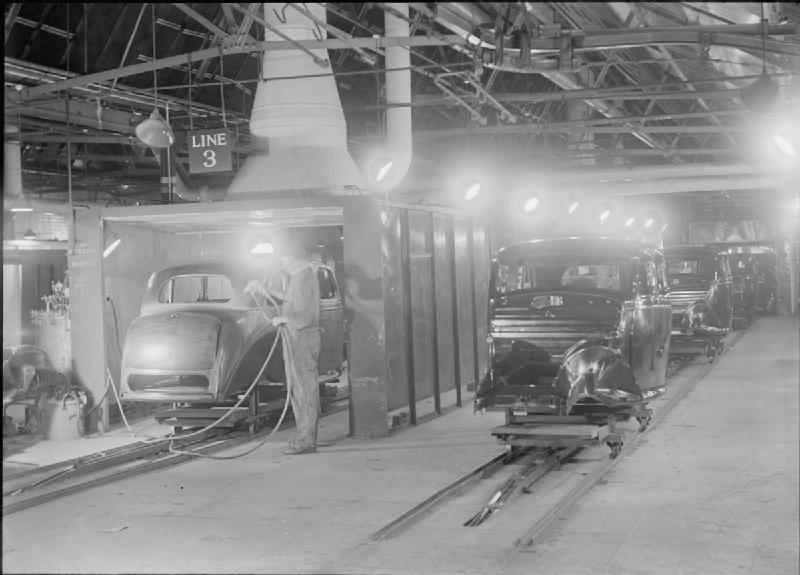 A general view of the paint shop at a car factory, somewhere in Britain. According to the original caption, the vehicle on the left in a spray booth "is starting on a 16-hour journey, will receive 3 coats of filler, 4 of primer, and 2 of cellulose, will pass through the long drying and hardening ovens, and return, like the bodies on the right, ready for body trimming and final assembly".