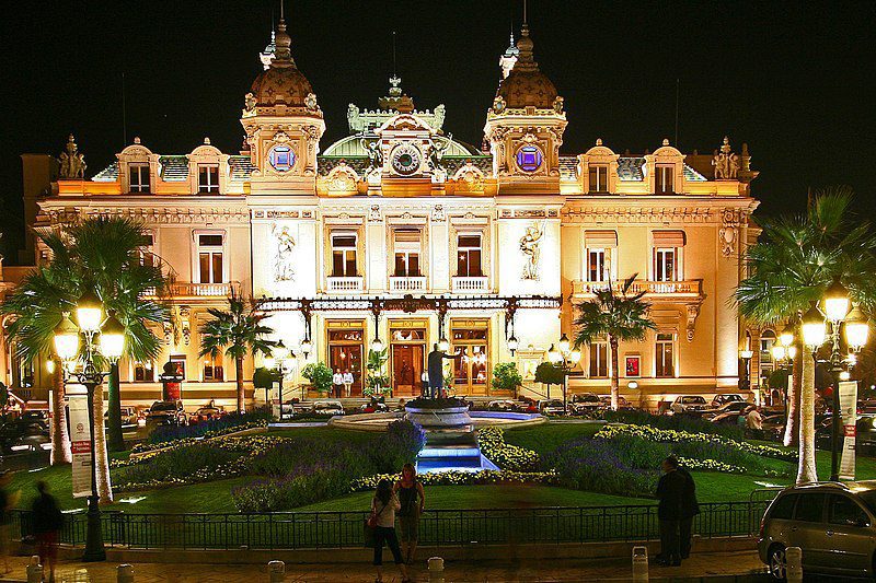 The Monte Carlo casino gleams with light against the dark sky, its Beaux Arts style facade glowing golden. Spotlights sweep over the building, illuminating intricate details on the balconies and pillars. 